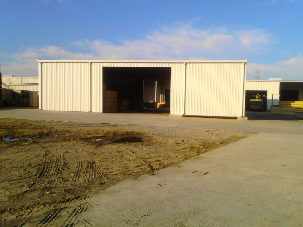 A large metal storage building with an open bay door sits on a concrete lot in an industrial yard.
