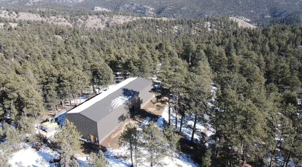 A steel warehouse surrounded by a forested mountain landscape with patches of snow on the ground and the building's roof.