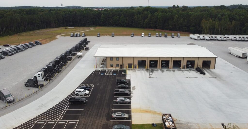 An aerial view of an auto shop building with multiple cars outside of it. The parking lot next to it has a variety of trucks.