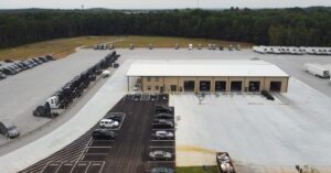 An aerial view of an auto shop building with multiple cars outside of it. The parking lot next to it has a variety of trucks.
