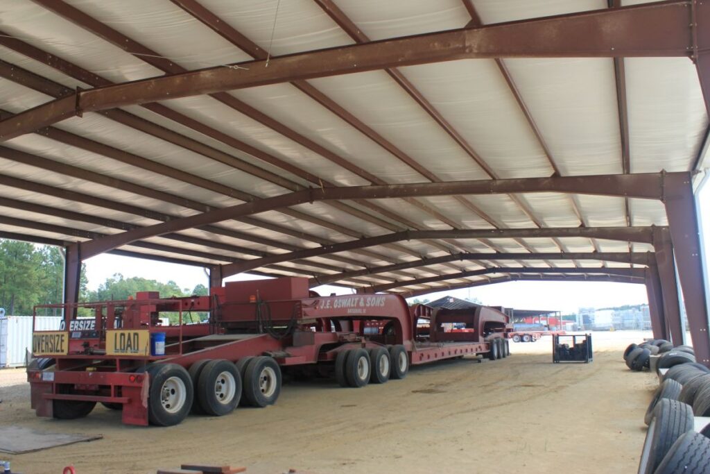 An oversized piece of farming machinery underneath a covered steel building with spare tires nearby.
