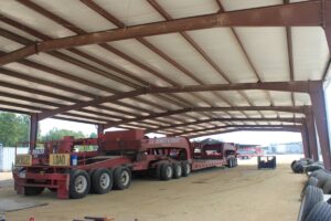 An oversized piece of farming machinery underneath a covered steel building with spare tires nearby.