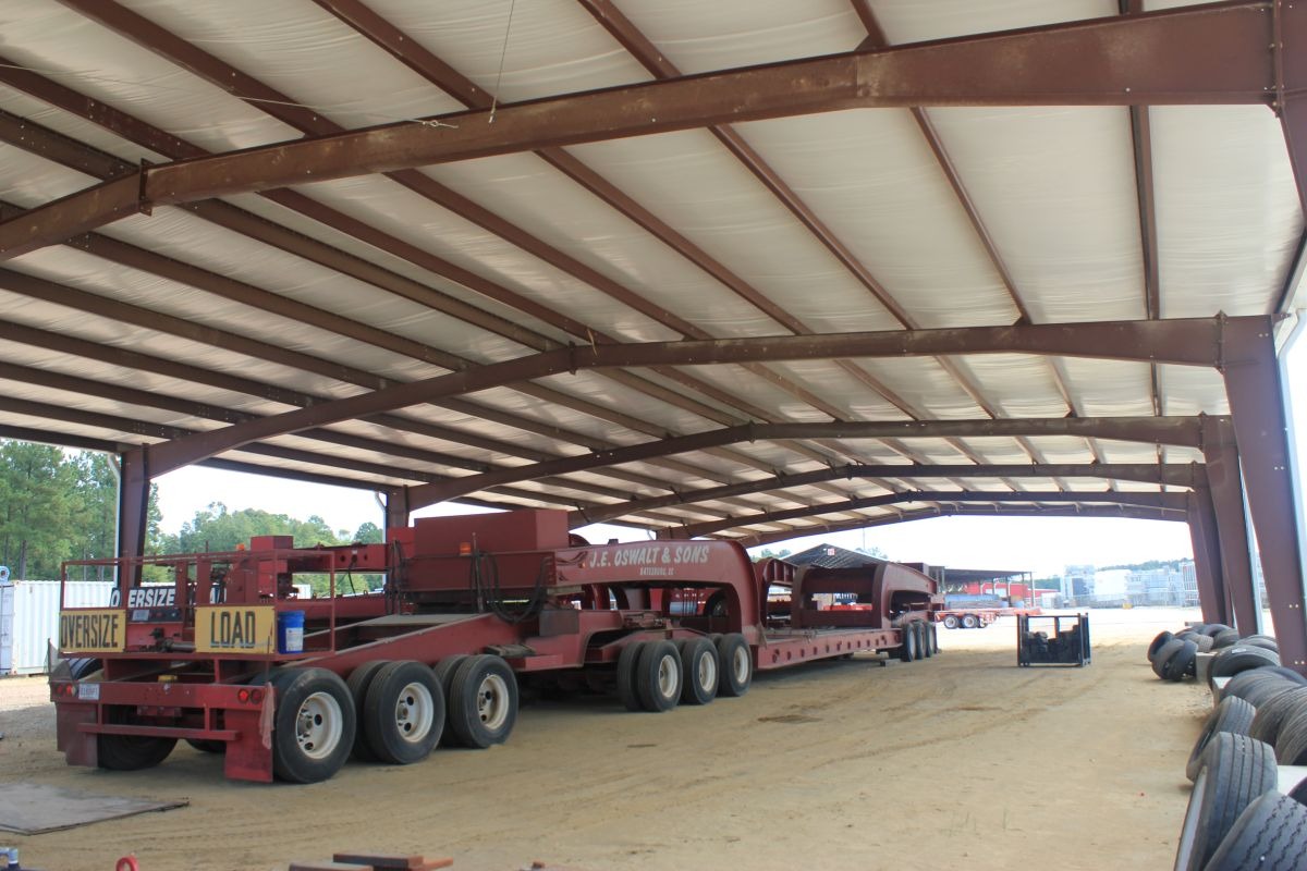 An oversized piece of farming machinery underneath a covered steel building with spare tires nearby.