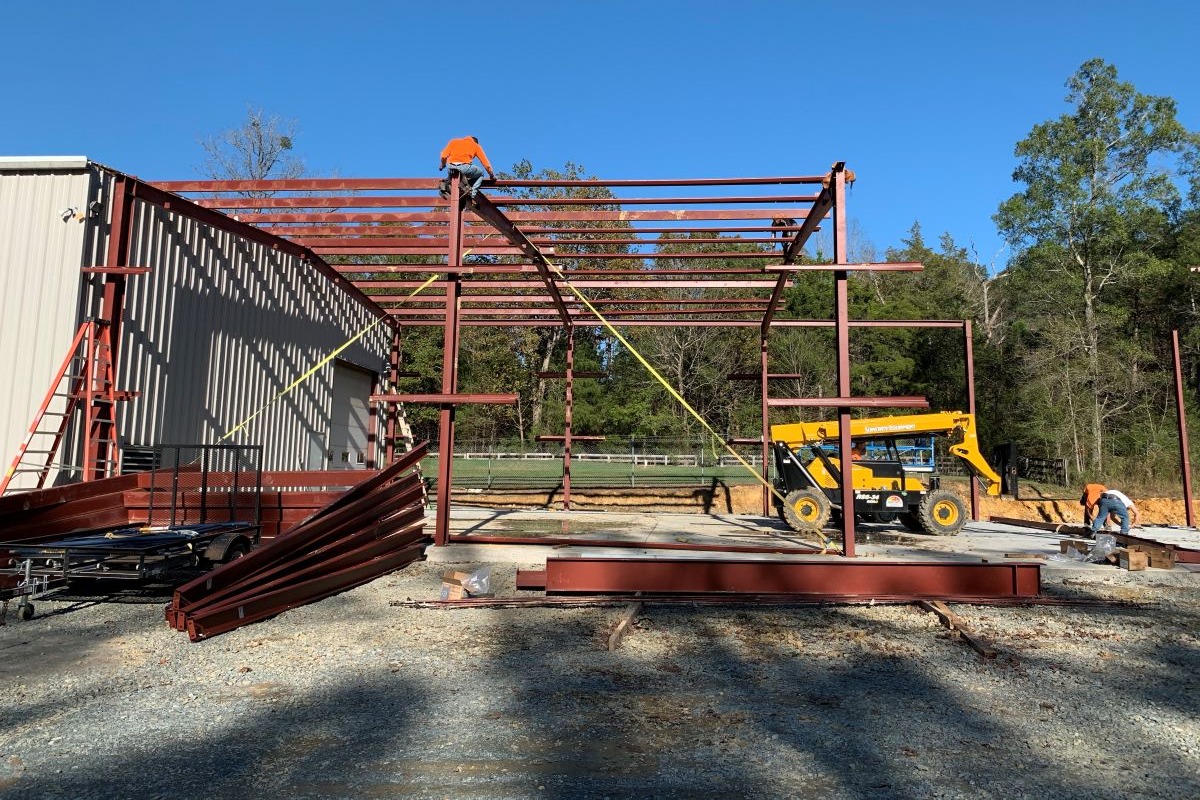 A large, red iron building being put together by workers. Once worker is on top of the structure creating the roof.