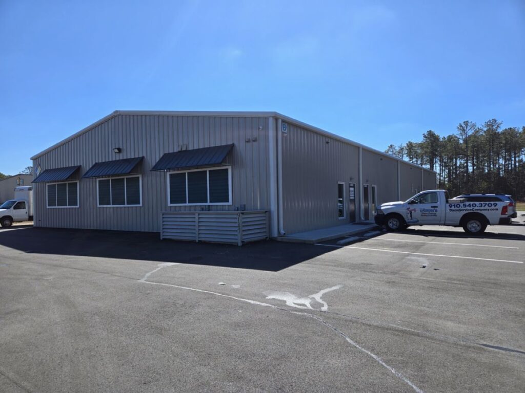 A large metal warehouse in the middle of a parking lot. There are several pickup trucks parked around the entrance.