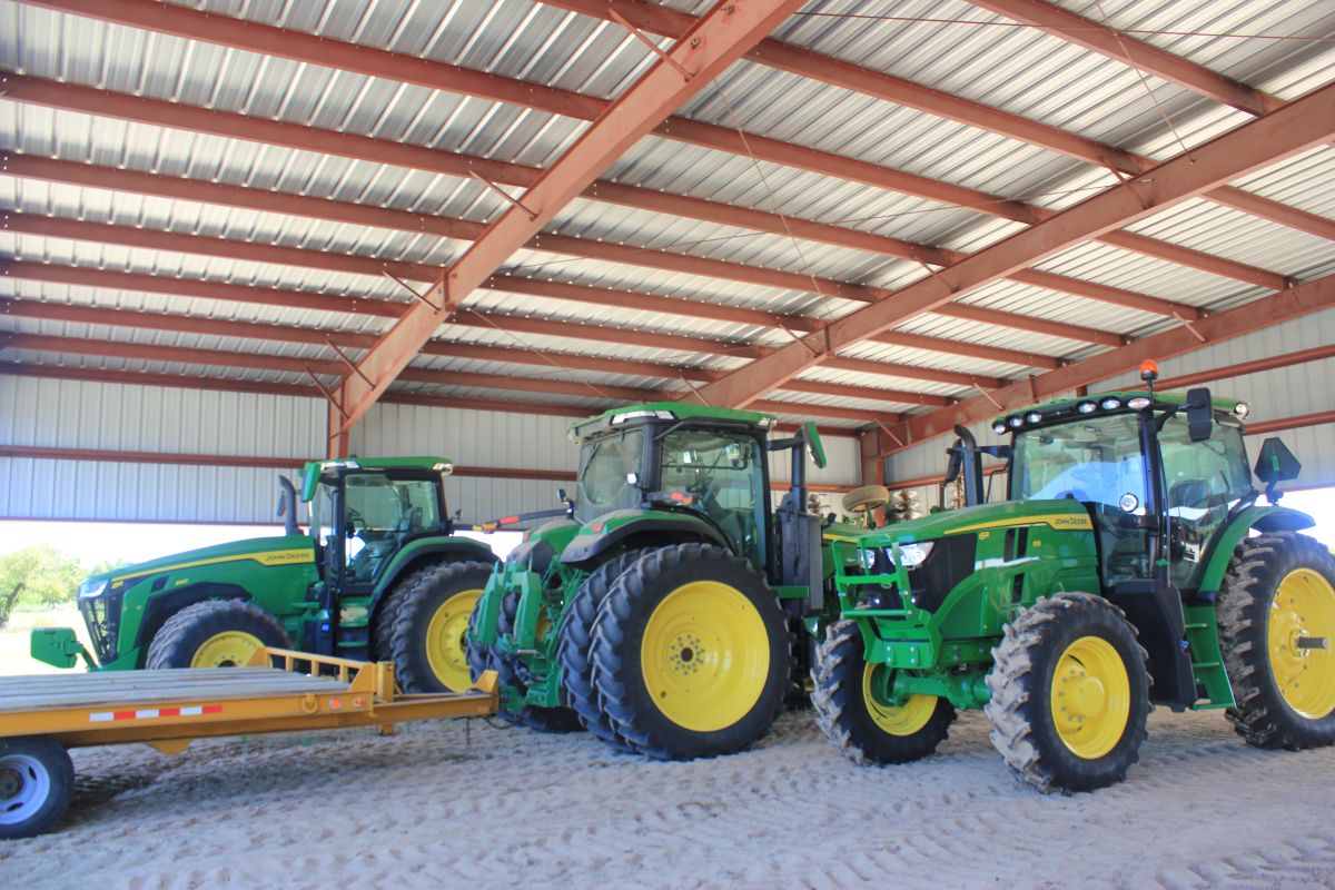 The interior of a barn with a steel roof. Inside the barn are multiple green tractors that are parked next to each other.